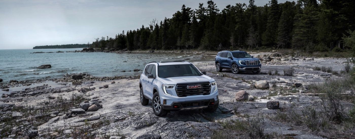 A white 2024 GMC Acadia AT4 and a blue Denali are shown near a lake after leaving a GMC dealer.