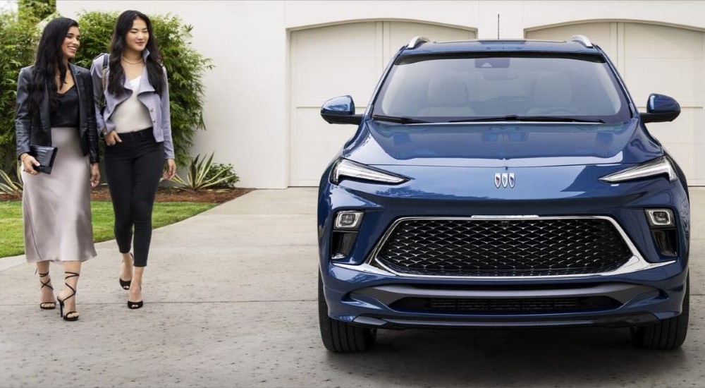 Two people are shown walking towards a blue 2024 Buick Encore GX Avenir parked in a driveway.